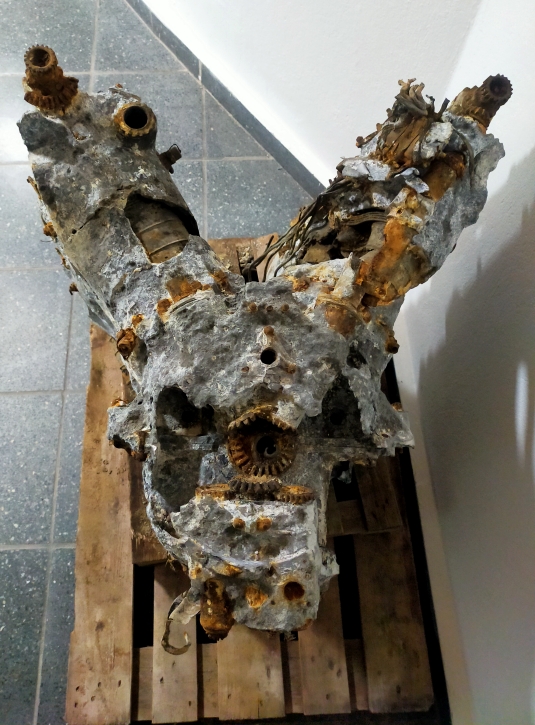 Die Draufsicht zeigt ein metallisches Objekt in Y-Form. Teile seiner Verkleidung fehlen. Es lagert auf einer Holzpalette in einem Innenraum